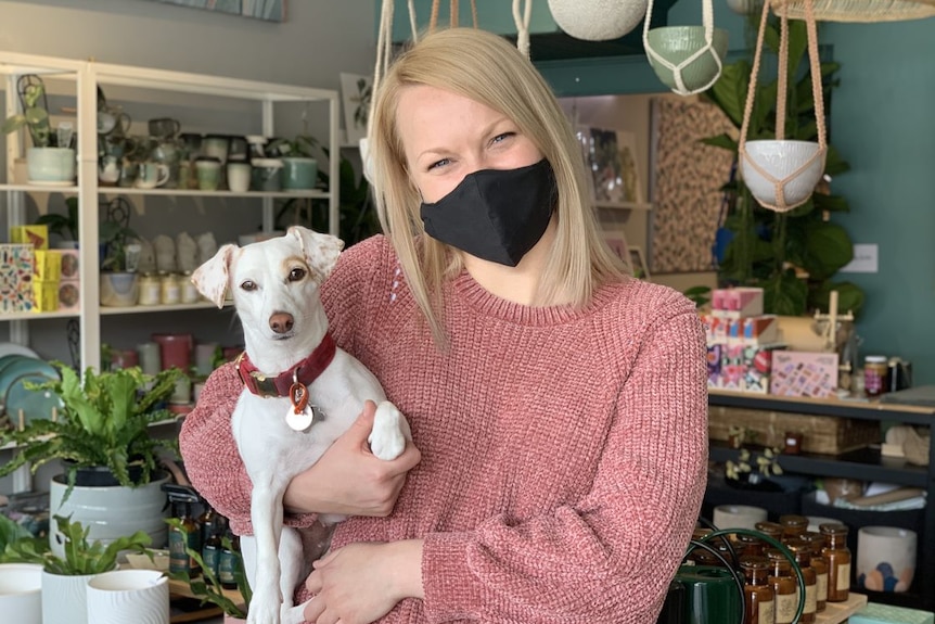 Bettina Tynan, wearing a mask, holds her dog Pip as she stands inside her gift store.