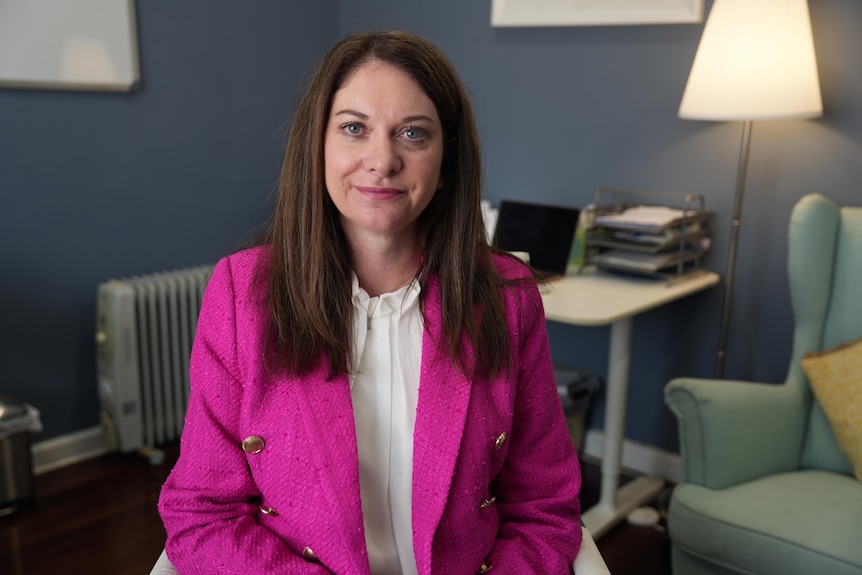A white woman with long brown hair wearing a pink blazer. She is sitting in an office