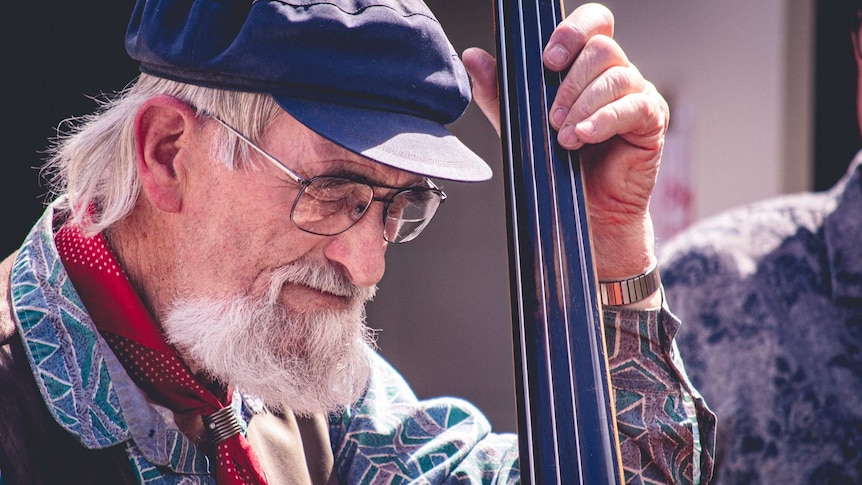 An elderly man plays the double bass with a subtle smile on his face