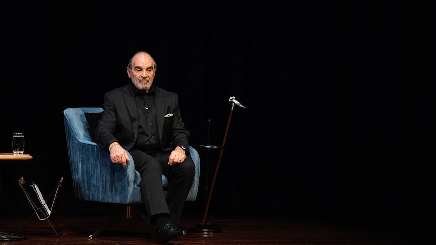David Suchet sits on a blue armchair on a wooden stage.