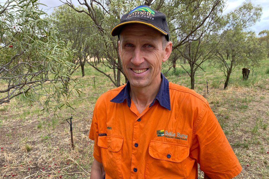 A man wearing a high-vis orange shirt and a blue cap smiling in front of some trees.
