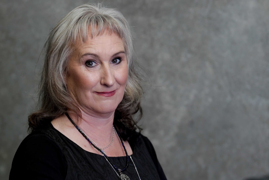 A middle-aged woman with grey hair and dark eyes stares at the camera.