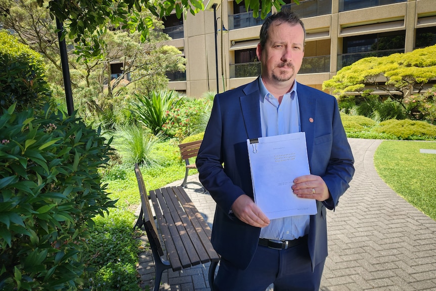 Man in suit standing NSW parliament courtyard holding an A4 document