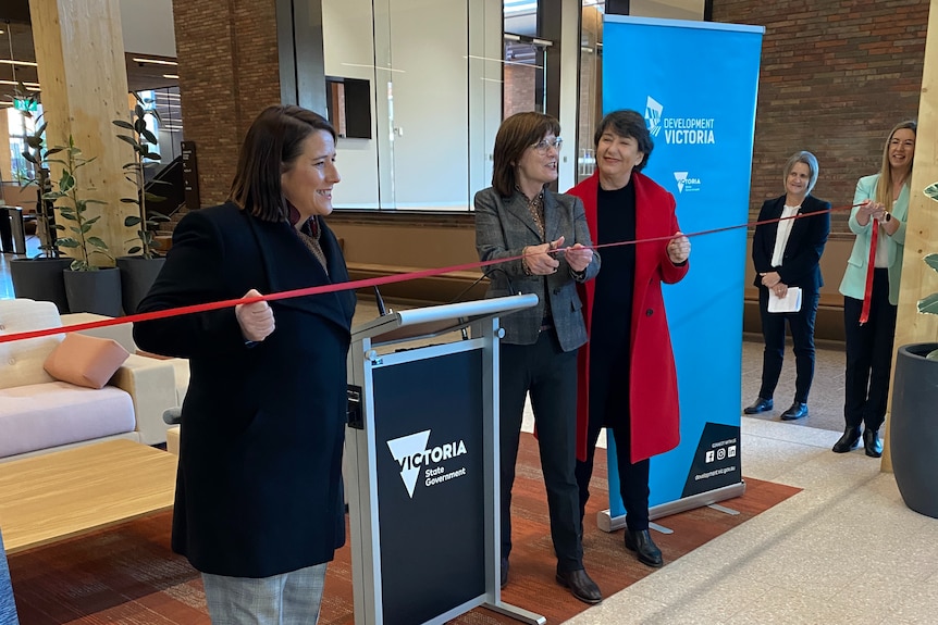 People watching as three women cut a red ribbon inside a new building.