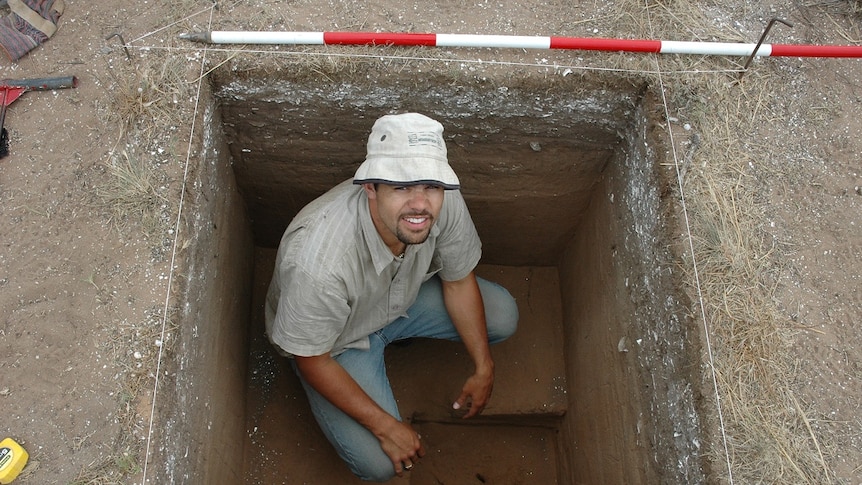 A man is squatting in a square dug out hole, looking up. There is a red and white pole near him.