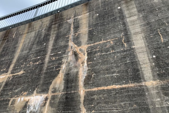 Cracks run toward the ground in a grey concrete dam wall