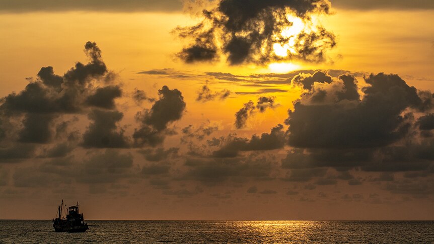 A fishing boat sails on calm waters as sun sets in cloudy golden-hued sky.