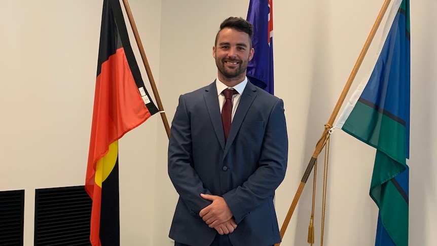 Former Councillor Ben Lucas is smiling and standing in front of the Aboriginal, Australian and Torres Strait flags.