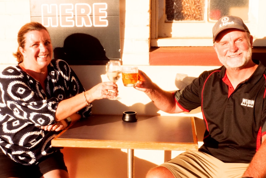 A woman and man sitting at a table clinking a wine and beer glass in cheers.