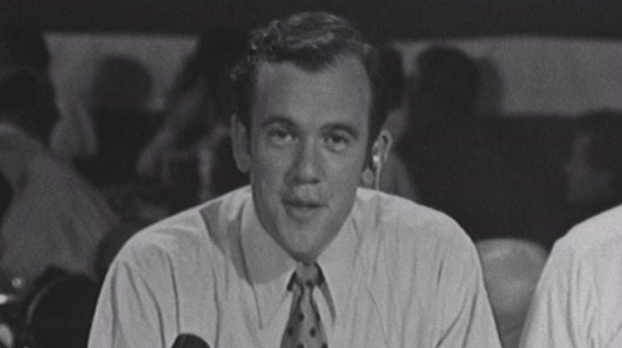 Mike Willesee looks into the camera, an earbud in his ear and a microphone on the desk.