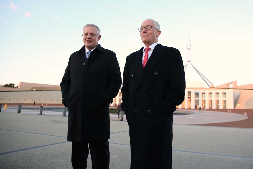 Two men stand with hands buried in warm coat pockets, Parliament House in the background. Malcolm Turnbull pulls a funny face.