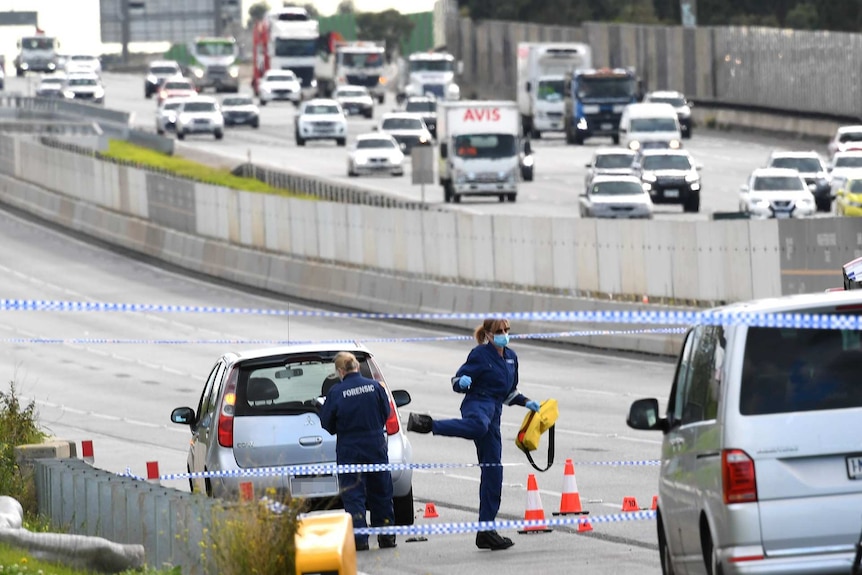 Two female police forensic officers examine a silver hatchback behind police tape on a freeway.