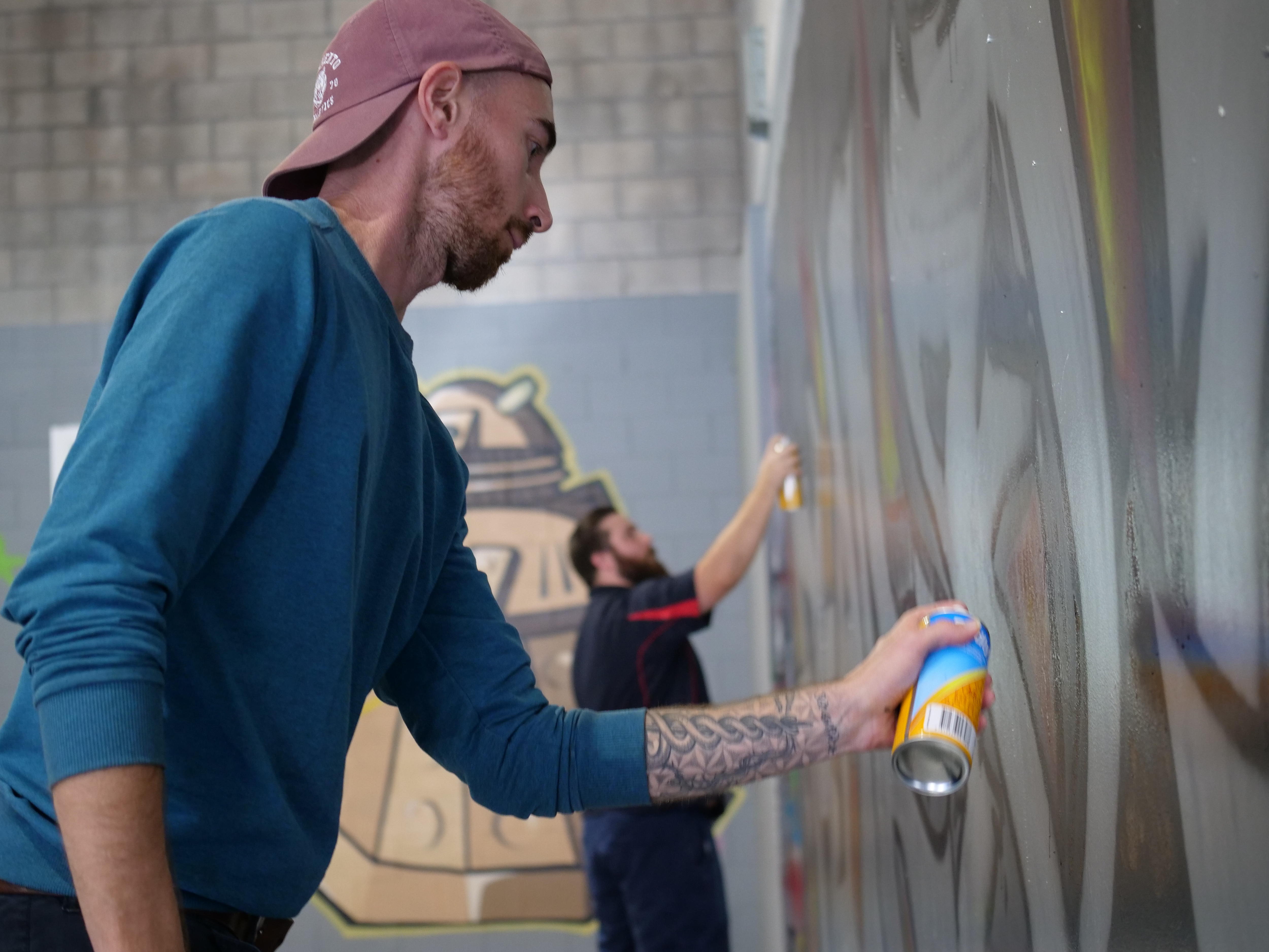 Two young men spraying a big graffiti piece onto an interior wall 