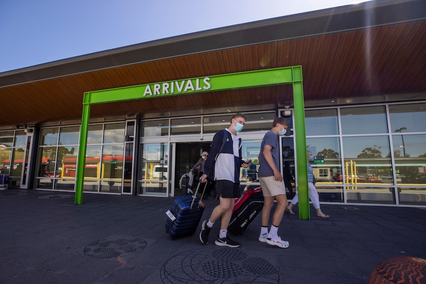 Two people in masks pull suitcases under a green arrivals sign 