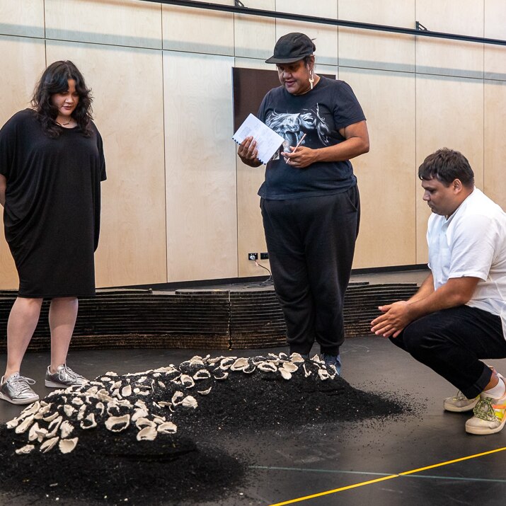 Shari Sebben, Elaine Crombie and Ian Michael gathered around a large mound of black glitter and shells that resemble a midden.