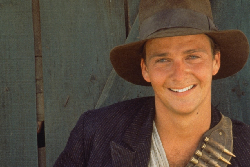 A young man smiles at the camera while wearing a fedora and a bandolier.