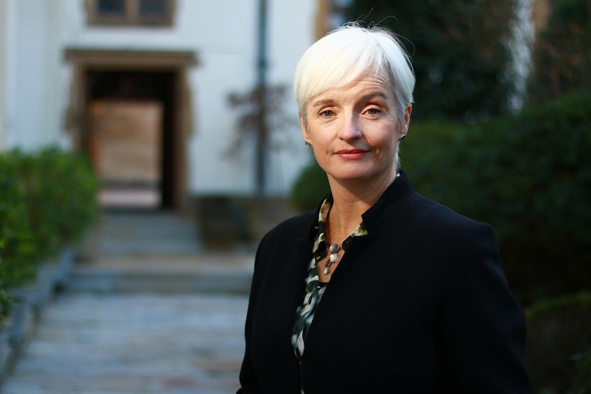 A white-haired woman in a dark suit jacket stands on a pathway into a blurry building in the distance