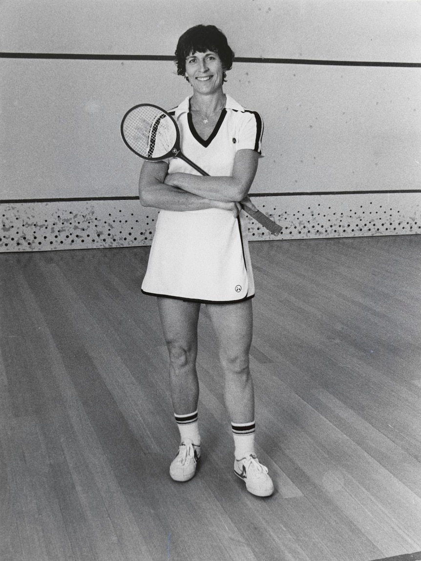 A woman stands on a squash court, smiling and holding a racquet.