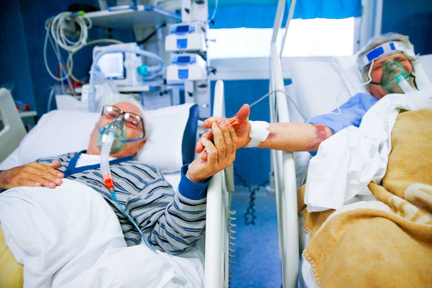 An elderly man and woman hold hands while lying in hospital beds