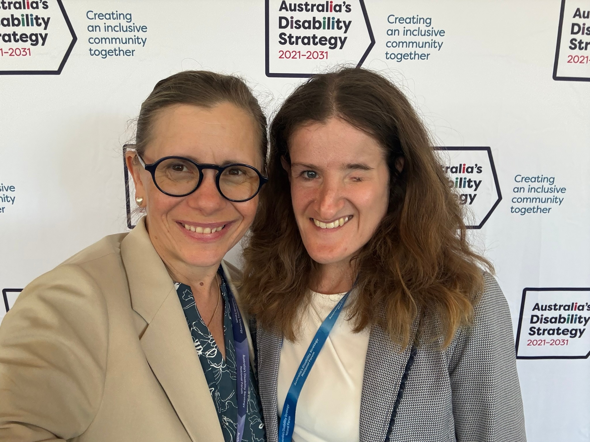 Two smiling, dark-haired women, one with facial difference, in front of a disability strategy backdrop.