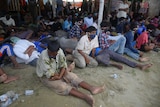 A large number of men sit and lie down on sandy ground.