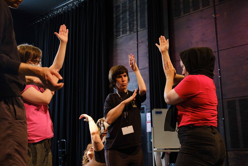 Sarah wears a black t-shirt and takes part in a theatre course with other people in a dark theatre room with her arms raised.