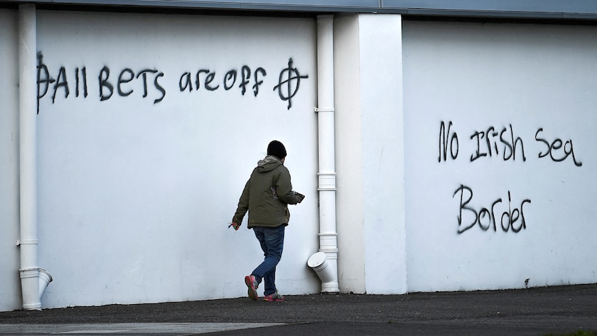 White wall with black graffiti that reads "all bets are off" and "no Irish sea border" with person walking past wall.