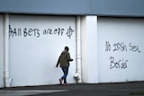 White wall with black graffiti that reads "all bets are off" and "no Irish sea border" with person walking past wall.