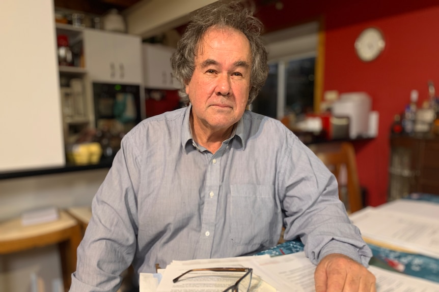 A middle-aged man is pictured at home at a desk with papers, looking serious.