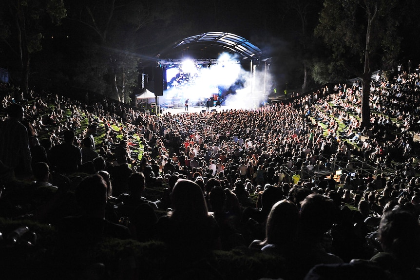 A sea of people seated, at night, in an ampitheatre in front of a bright stage seen in the distance