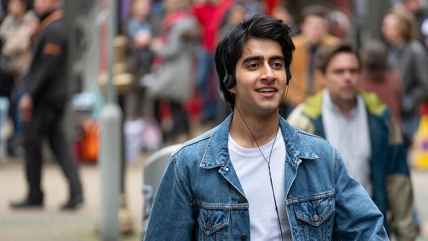 Viveik Kalra walks down the street listening to music on overhead headphones, dressed in white t-shirt and blue denim jacket.