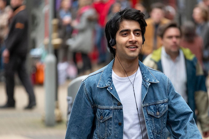 Viveik Kalra walks down the street listening to music on overhead headphones, dressed in white t-shirt and blue denim jacket.