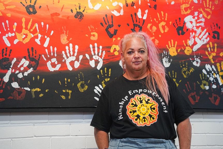 Head to waist shot of Lisa standing in front of an Indigenous painting with hand prints. 