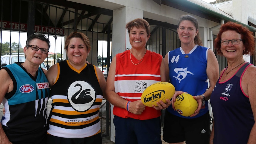 Five women in different football jumpers pose for a photo outside Fremantle Oval.