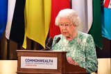 Queen Elizabeth II speaks during the formal opening of CHOGM. She is at a podium in front of several Commonwealth nation flags.