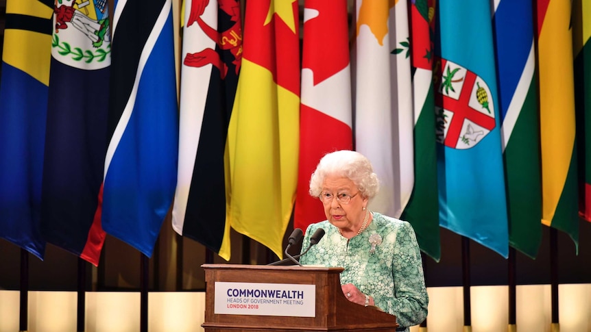 Queen Elizabeth II speaks during the formal opening of CHOGM. She is at a podium in front of several Commonwealth nation flags.