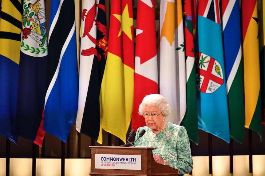 Queen Elizabeth II speaks during the formal opening of CHOGM. She is at a podium in front of several Commonwealth nation flags.