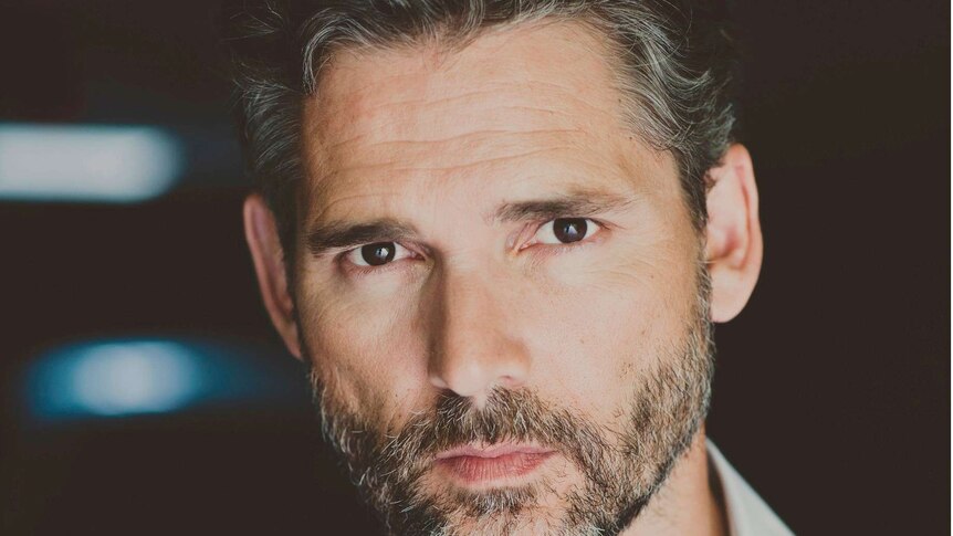 A middle-aged man with greying hair stares into a camera with a black background (Eric Bana)