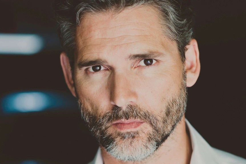 A middle-aged man with greying hair stares into a camera with a black background (Eric Bana)