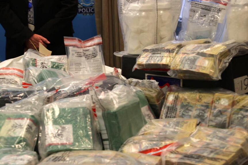 Drugs and cash are displayed on a table in evidence bags as police stand in the background.