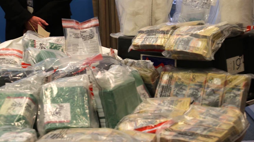 Drugs and cash are displayed on a table in evidence bags as police stand in the background.