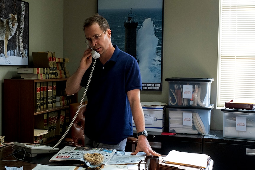 A man with glasses and dark blue polo shirt stands at desk covered in files with phone raised to ear in office space.