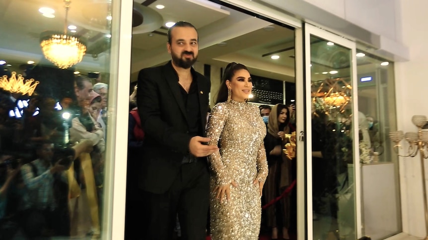 A woman in a sparkling silver dress and a smartly dressed man enter a room. In the reflection of glass there are photographers