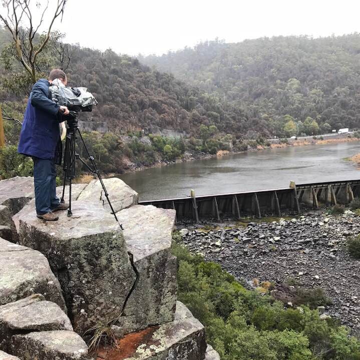 Cameraman standing on rock filming water about to spill over dam wall in background.