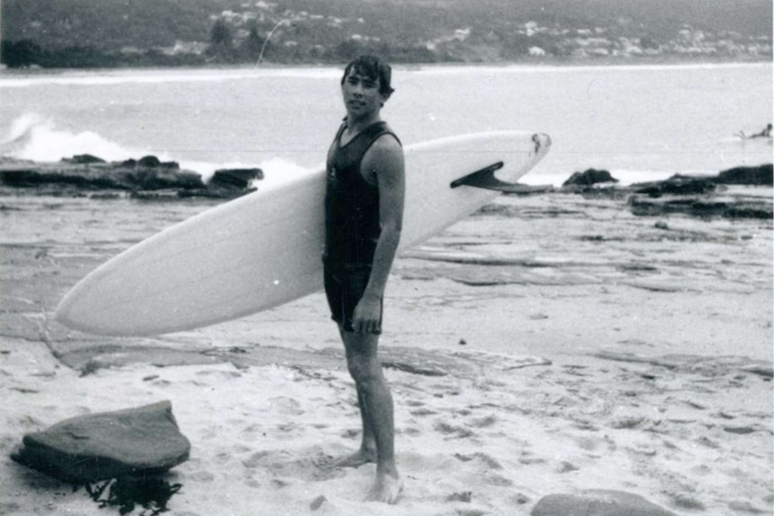 Black and white photo of a teenaged male wearing a wet suit and hoding a surfboard at the beach.