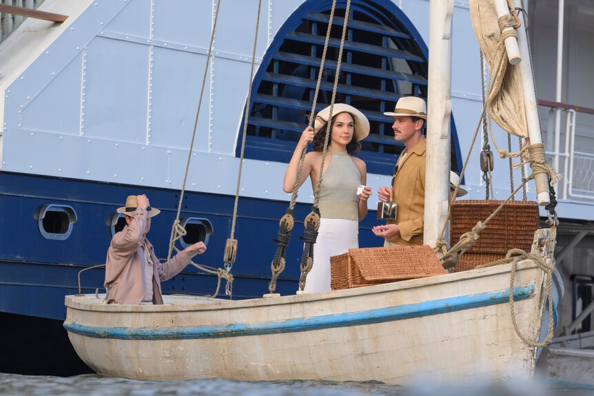 A couple aged in their 30s, wearing light linen clothing and hats, stand together on a dingy, attached to a large ship