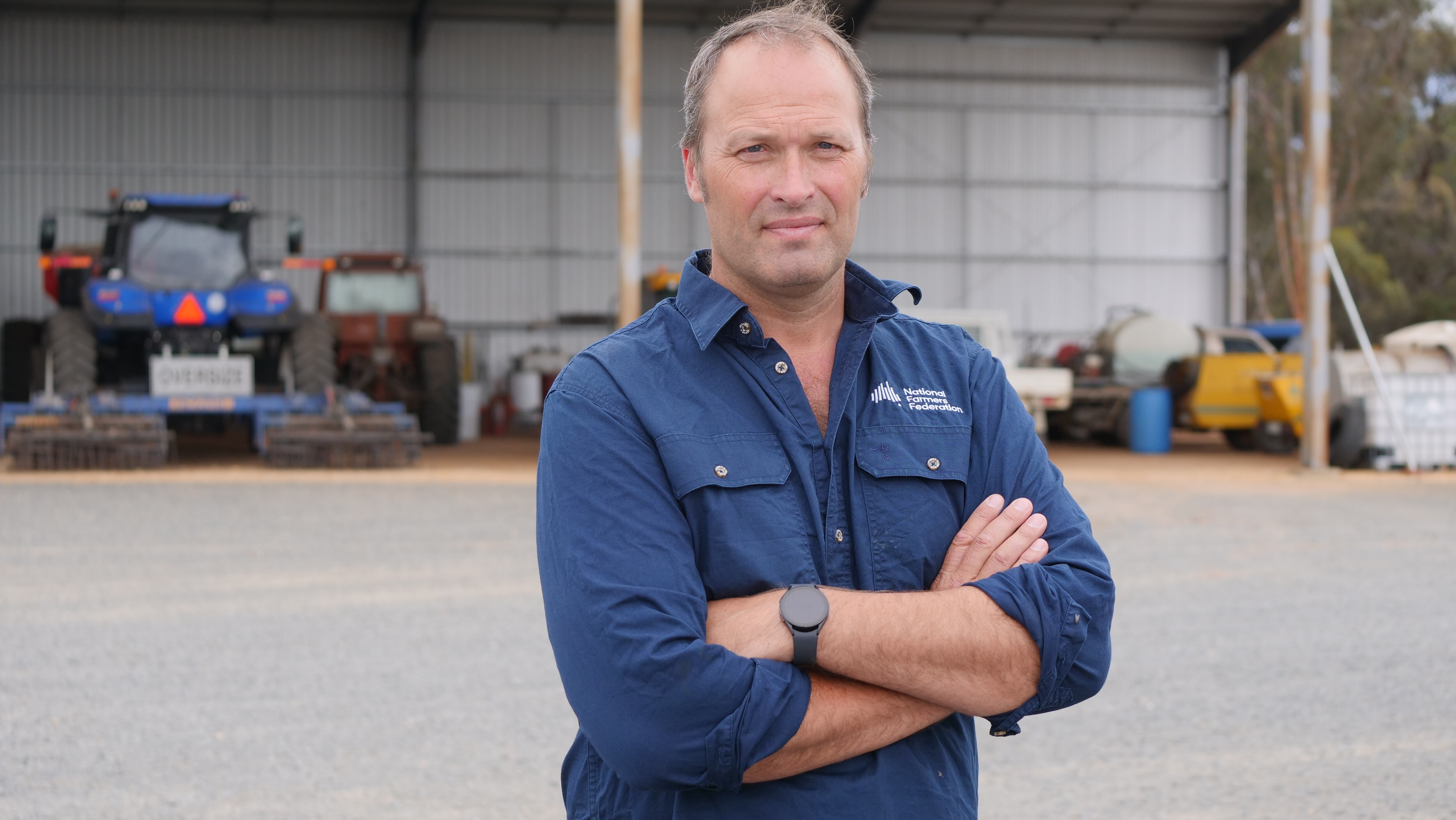 A tanned man wearing a dark blue work shirt crossing his arms. In the background there are tractors and farm equipment.