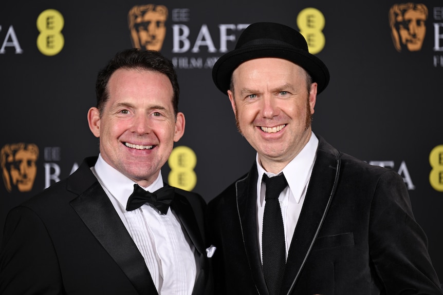 Mid close up of two men in suits, smiling on the red carpet.