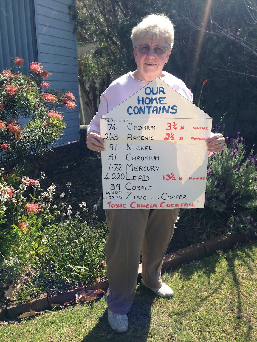 A woman with grey hair standing in a garden holding up a sign.