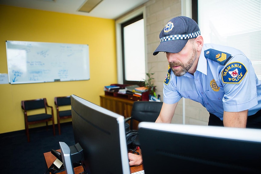 A policeman in peaked cap leans in to two computer screens to scrutinise information.
